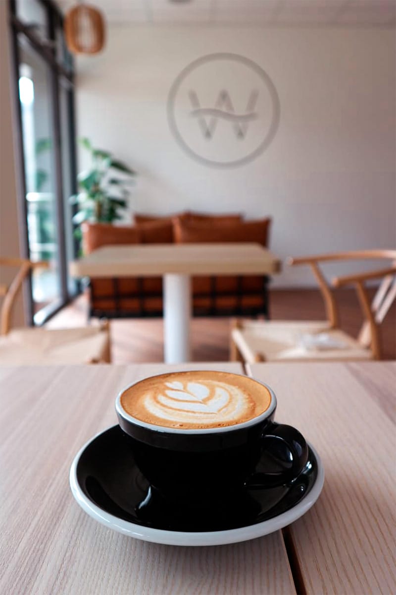Photo of a latte inside Way Coffee with the logo on the wall in the background.